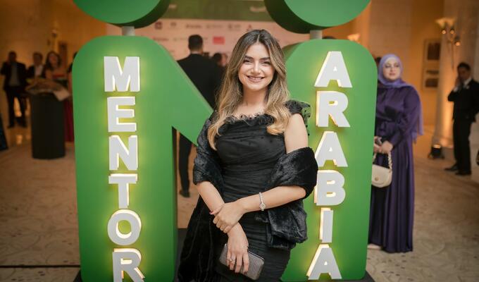 Jordanian woman in formal wear stands in front of green Mentor Arabia sign