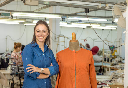 A smiling young woman standing with arms folded beside a dressmaker mannequin wearing a bright orange shirt. The background is of a factory, with women and clothing on racks.