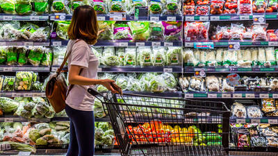 Woman facing a wall of refrigerated produce, with shopping trolley and holding a mobile phone.
