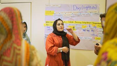 Pakistani woman in head scarf speaks to workshop, standing in front of white board
