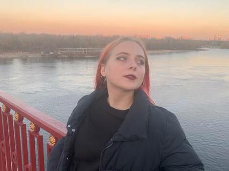 Woman with pink hair wears black jacket while leaning on bridge railing