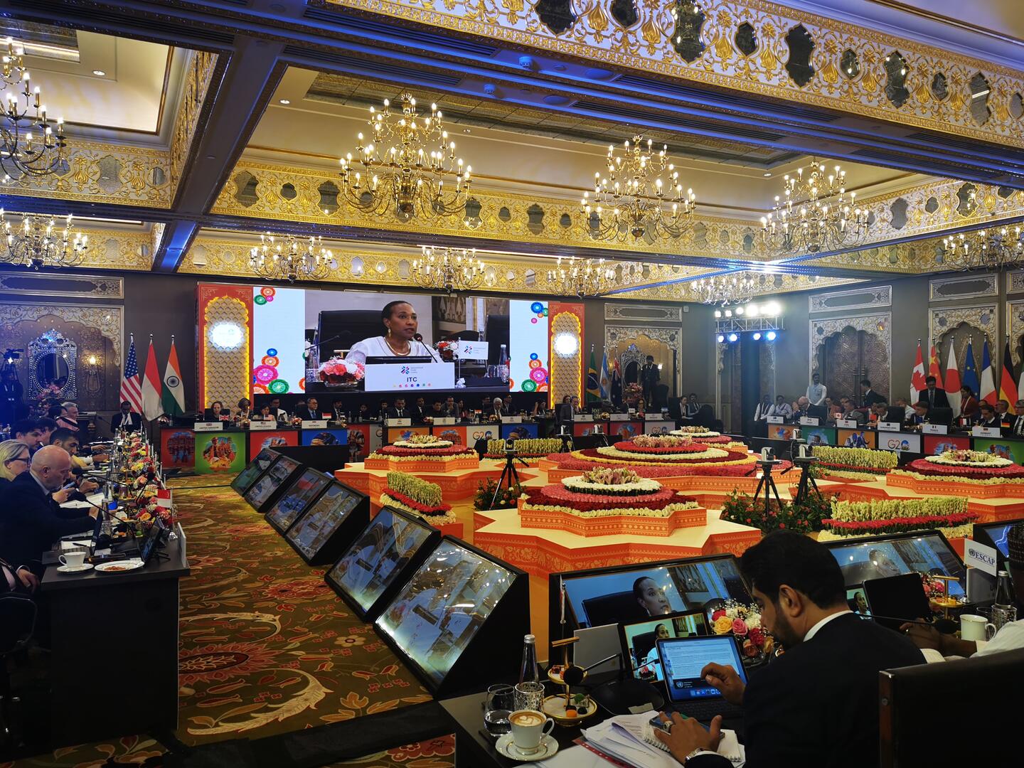 ITC executive director Pamela Coke-Hamilton addressing G20 ministers on a large projector screen, with the ministers shown seated around the perimeter at tables facing inwards, with a large floral display in centre of room.