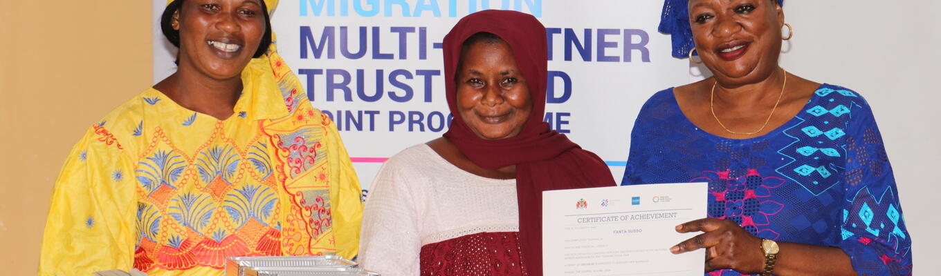Three Gambian women in bright clothing hold certificates and goods received from a grant