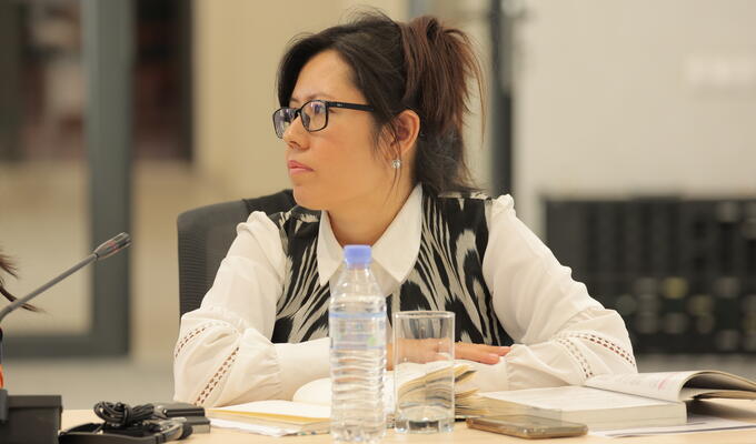 Woman in glasses sits at table with arms crossed, listening to presentation