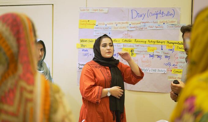 Pakistani woman in head scarf speaks to workshop, standing in front of white board