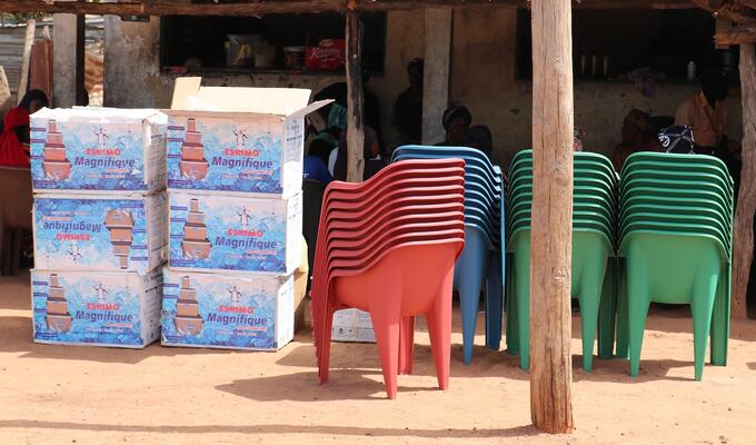 Bright red and green plastic chairs are stacked next to cardboard boxes in a Gambian market