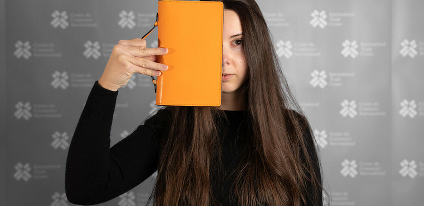 Young, serious-looking woman with long dark hair in a black shirt, against a grey ITC background, holding an orange notebook over half of her face lengthways