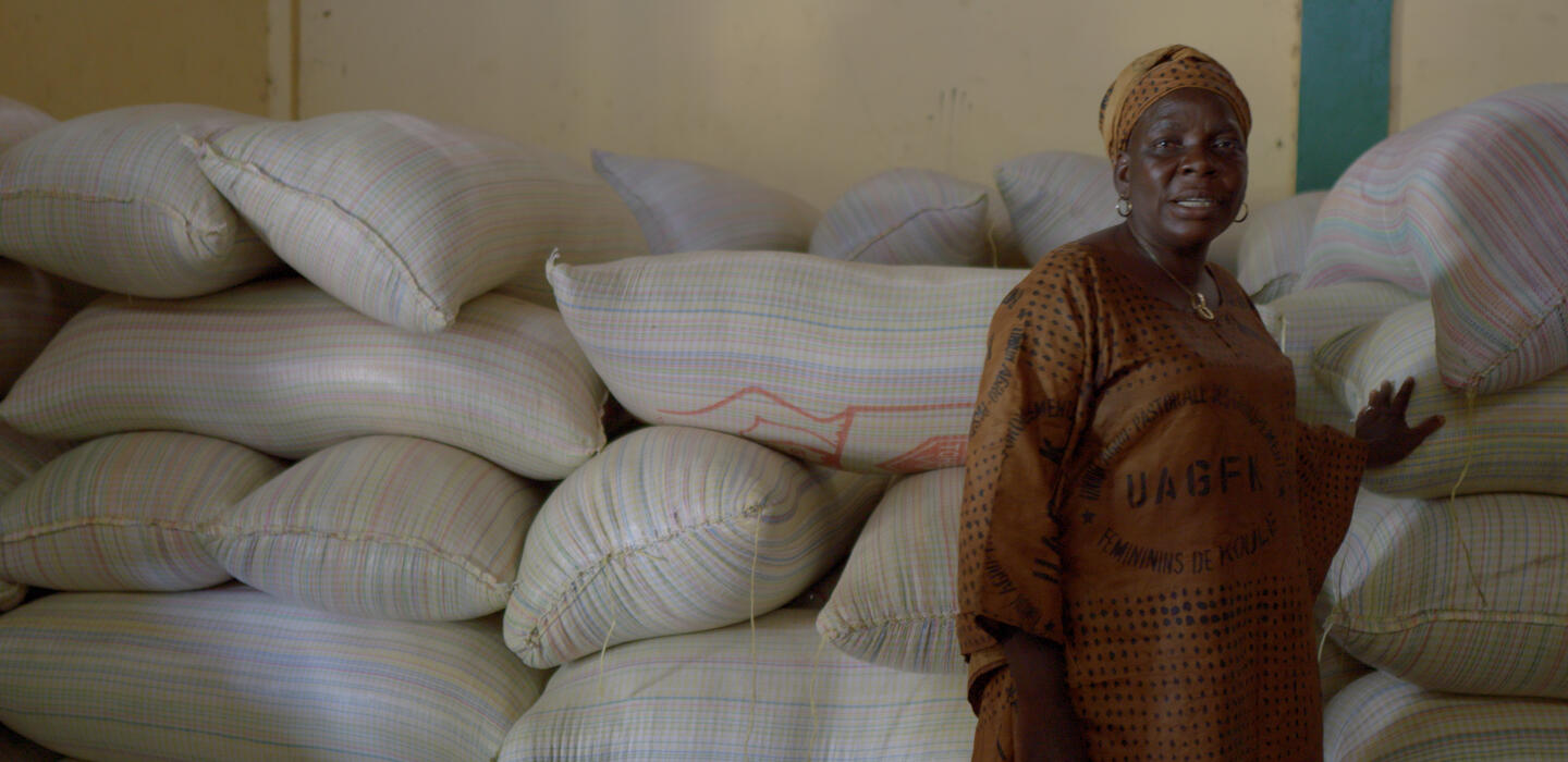 Guinean woman in a brown dress walks past sacks of rice. 