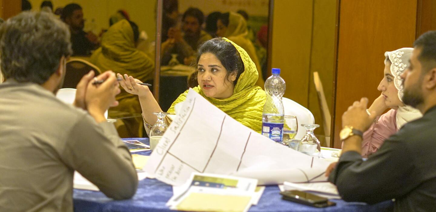 Woman in yellow headscarf speaks to group at table covered with notes