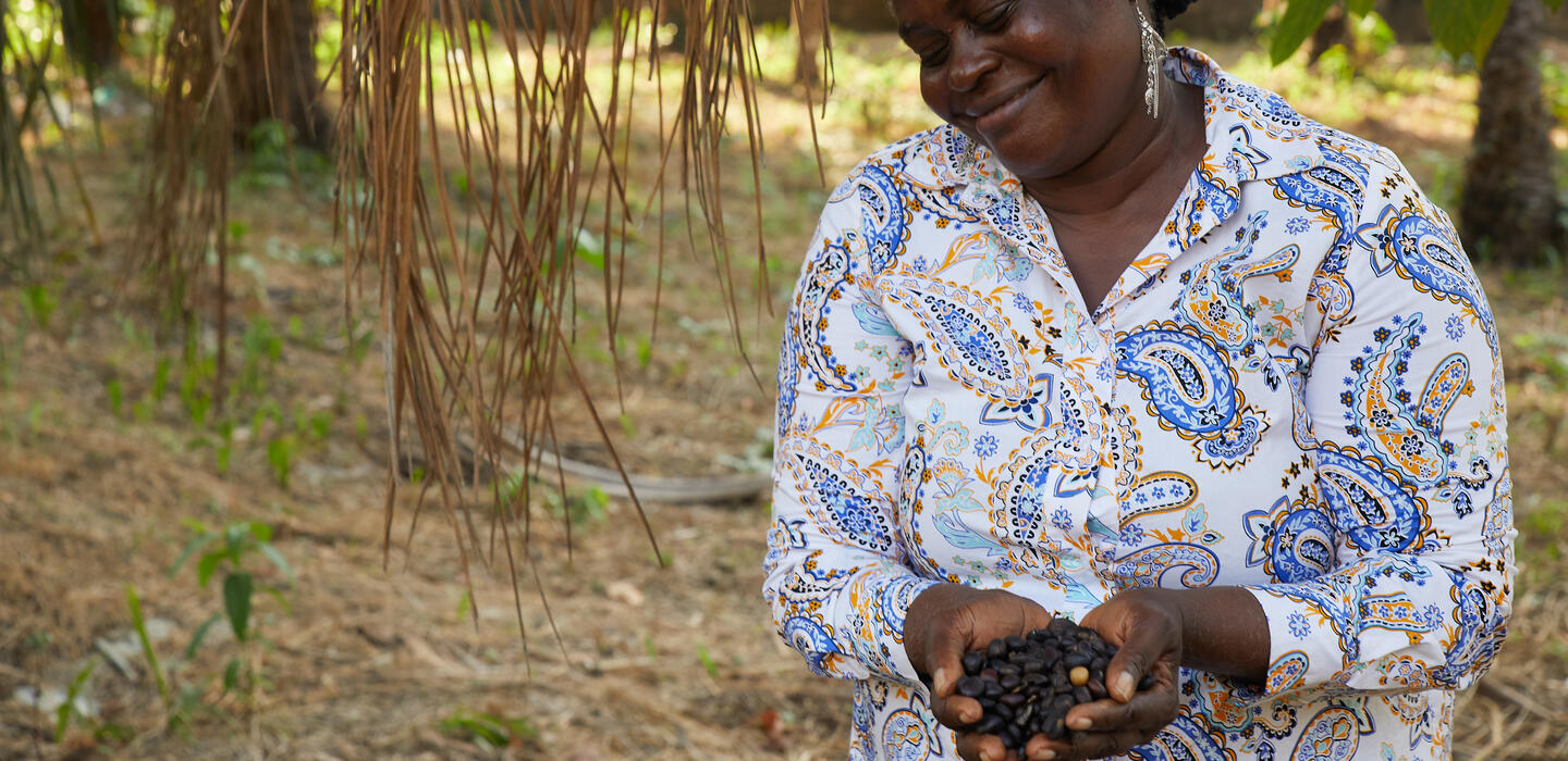 Au cœur d'une plantation, une femme, paumes ouvertes, présente des noyaux de fruits de palmier.