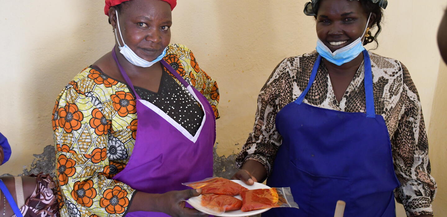 Two South Sudanese women wearing aprons and masks are holding food packets.