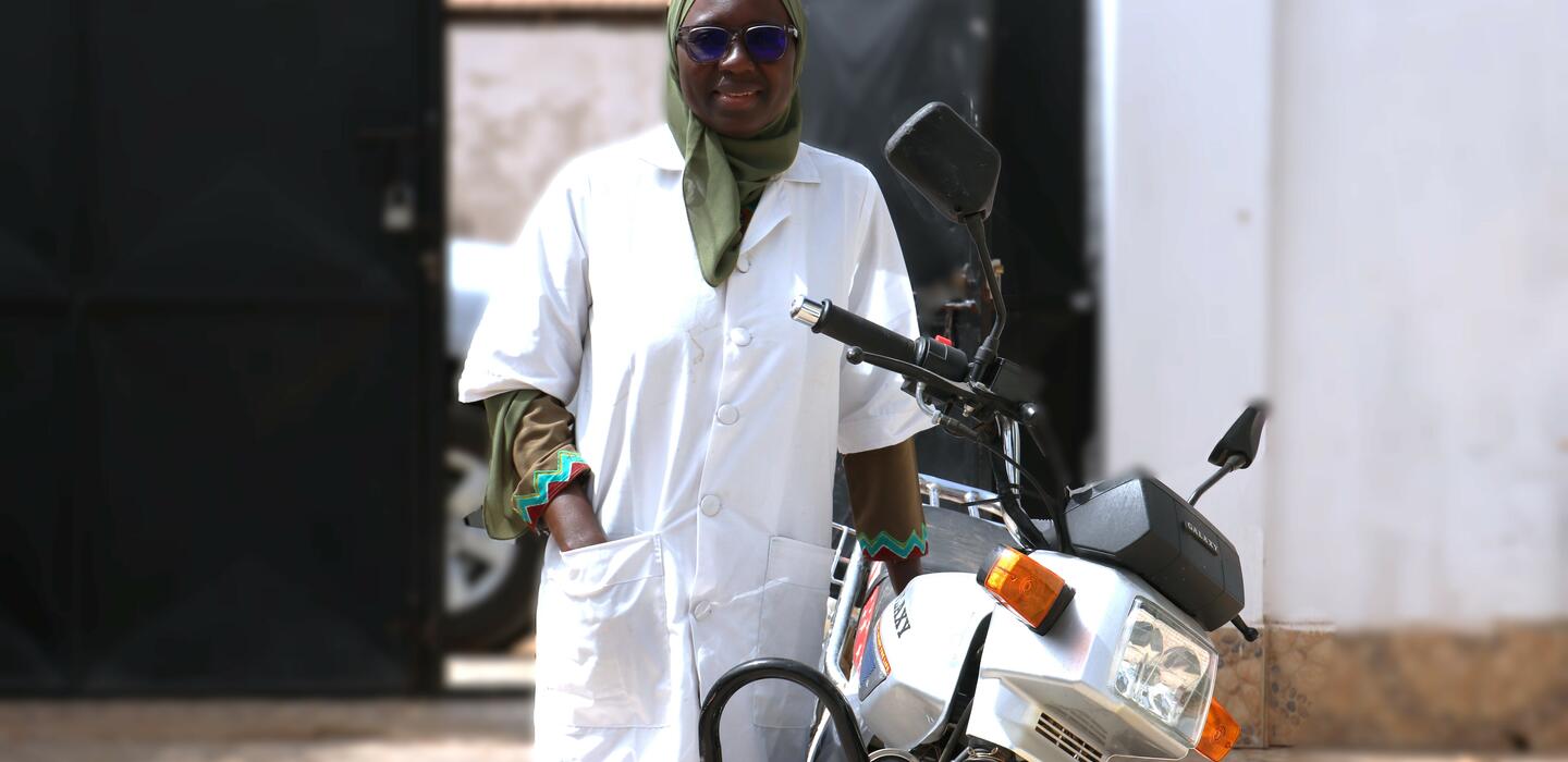 Gambian woman in white top and green headscarf stands next to motorcycle