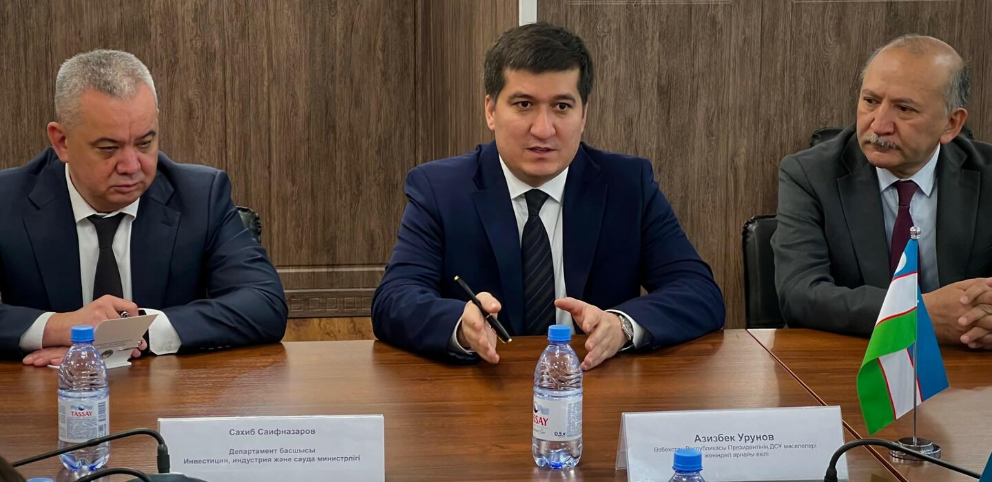 Three men in dark business suits sit at wooden table with small Uzbek flag