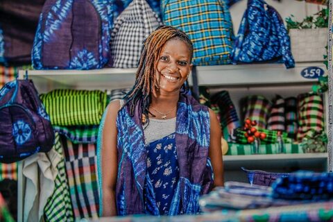 Gambian woman smiling at the camera in front of a wall of colourful fabric bags behind her on shelves
