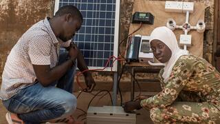Man and woman crouching with a small solar panel and its equipment in center of picture.