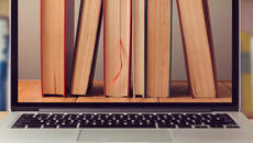 A laptop on a table which has a desktop image of standing books showing the pages. The background is of a shelf of books.