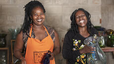 Two women of African descent with big smiles, one of them wearing an orange apron. There are low shelves of glass vases behind them along a brick wall, and one woman is holding a large glass vase